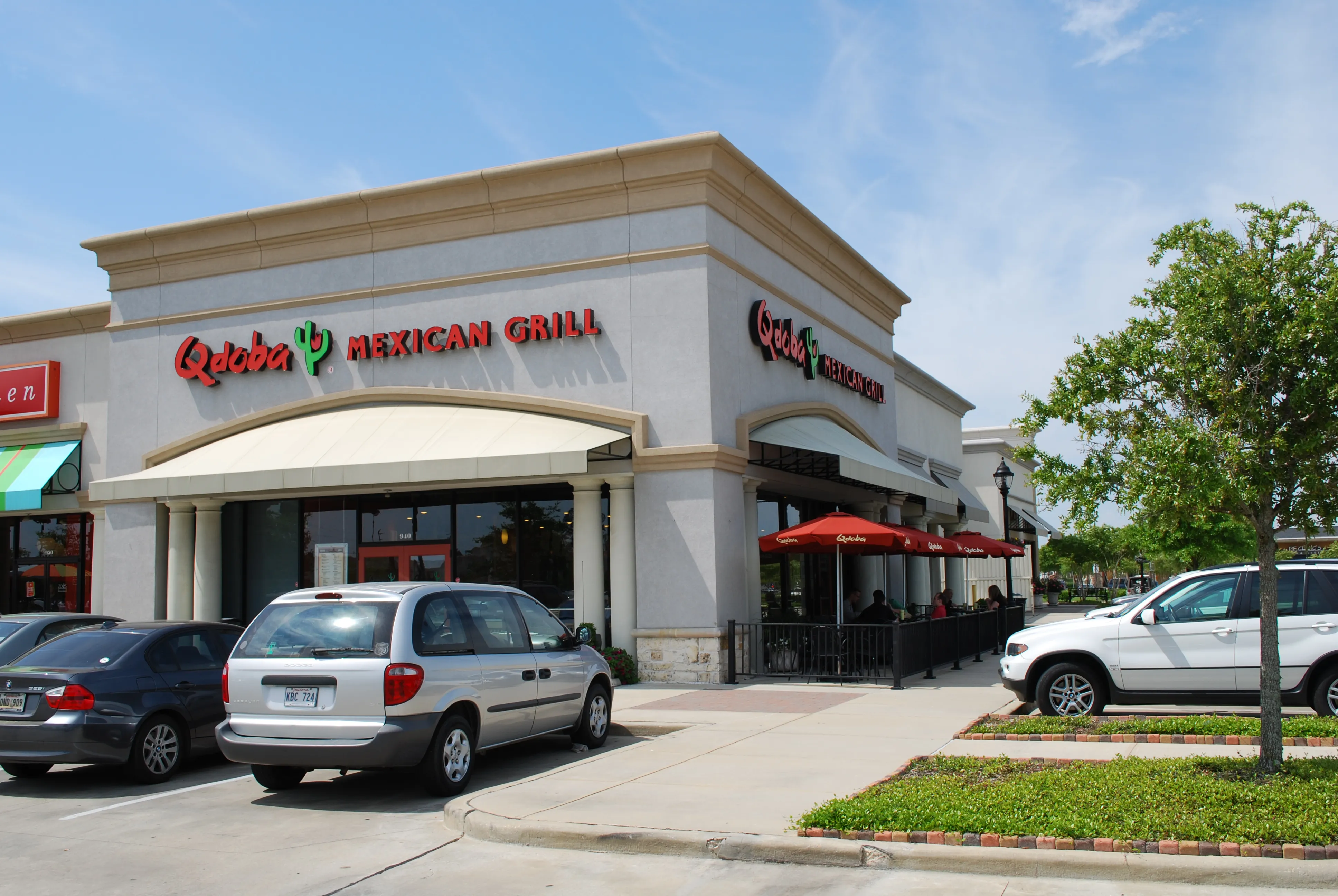 Corner view of the Qdoba Mexican Grill showing the wrap-around signage and red patio umbrellas.